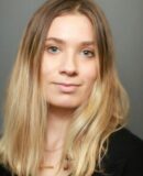 Emily_headshot A headshot of a person with shoulder-length blonde hair, neutral expression, and a dark top, set against a plain background.