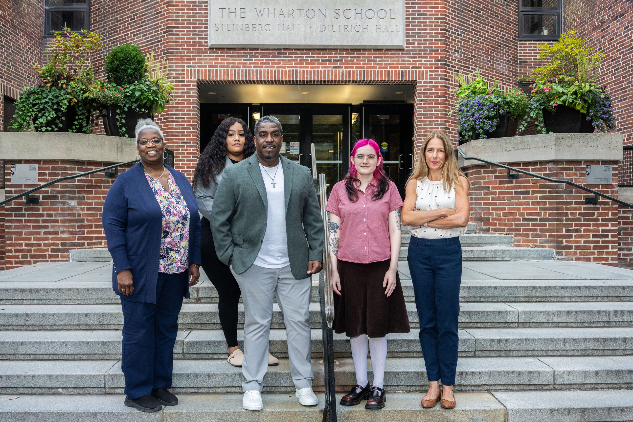 A small group of people standing on the steps in front of The Wharton School, Steinberg Hall. The background features a brick building with potted plants.